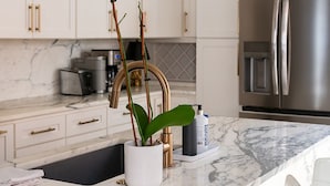 Una cocina moderna con gabinetes blancos, encimera de mármol, refrigerador de acero inoxidable y una lámpara colgante.