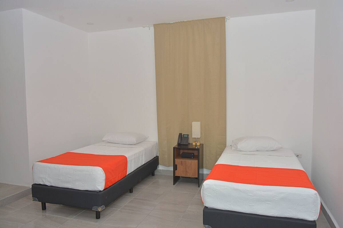 Une chambre d’hôtel avec deux lits simples, chacun agrémenté de draps blancs et de touches de couleur orange, une petite table de chevet en bois avec une lampe, et un rideau beige.