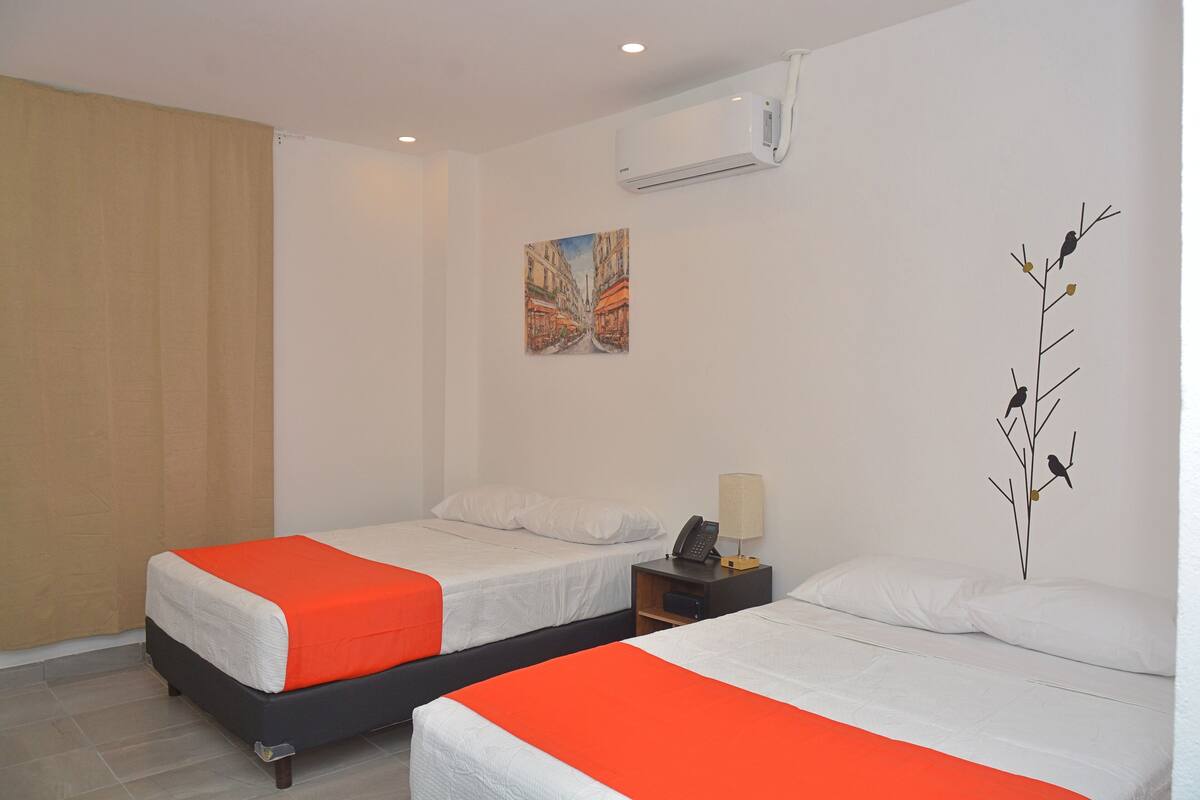 Une chambre d’hôtel avec deux lits, chacun agrémenté de draps blancs et de touches de couleur orange, une table de chevet avec une lampe, un tableau au mur et un appareil de climatisation.