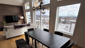 Un salon moderne avec une table à manger en bois foncé, des bancs et un canapé. Un téléviseur à écran plat est fixé au mur, et de grandes fenêtres offrent une vue sur un paysage enneigé.
