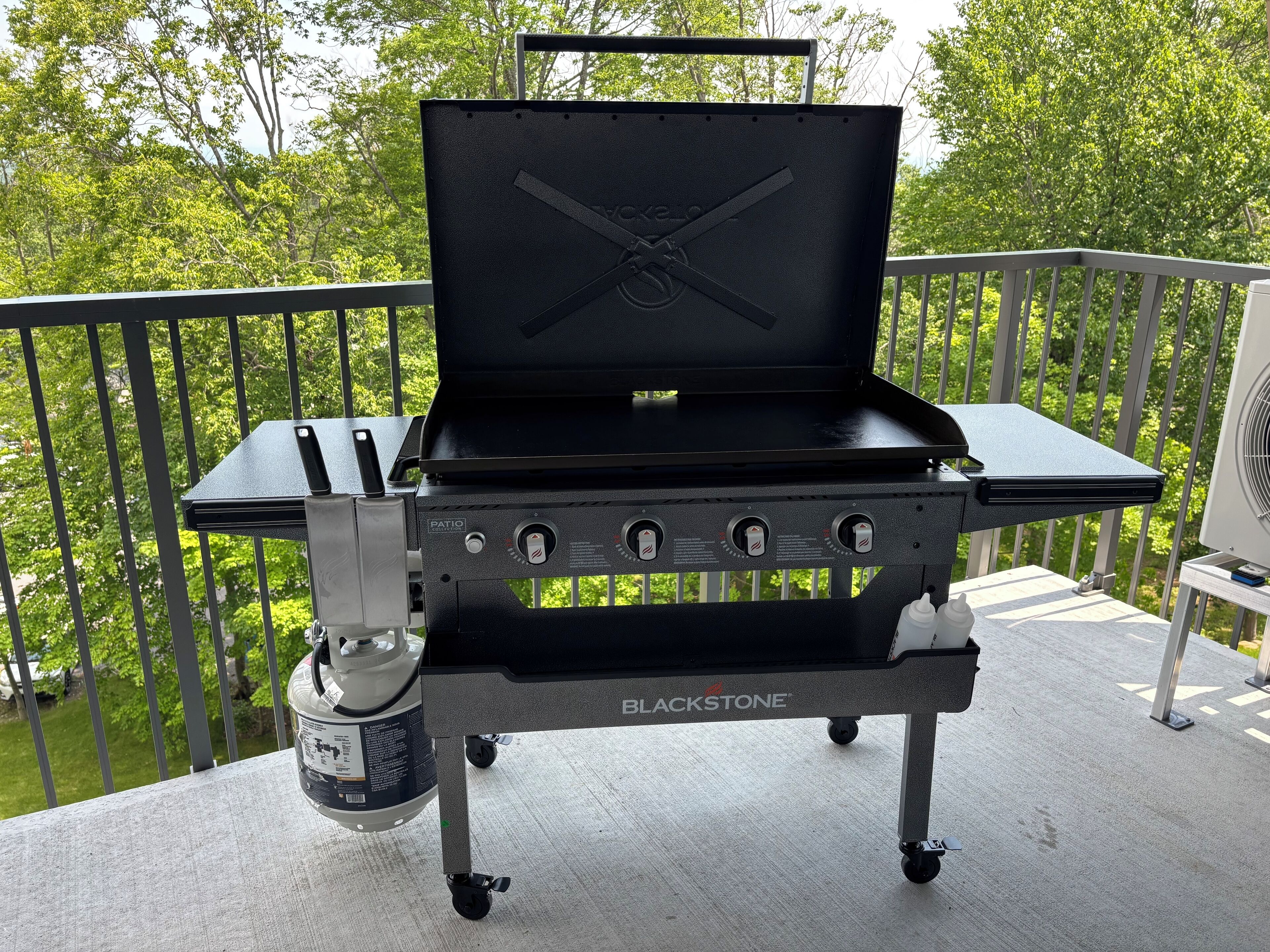 Una parrilla portátil Blackstone para el patio con cuatro quemadores, un quemador lateral y una tapa, en un balcón con barandilla y un ventilador al fondo.