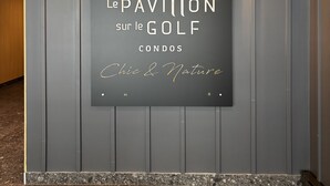 Une enseigne portant l’inscription « Le Pavillon, résidences de luxe avec vue sur le terrain de golf, chic et nature », fixée sur un mur sombre, avec deux lampes murales.