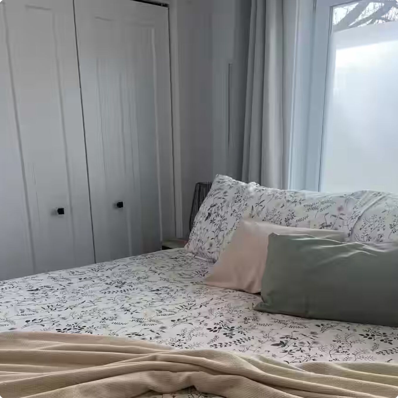 A bedroom with a bed, floral-patterned bedding, a beige blanket, and a window with curtains.