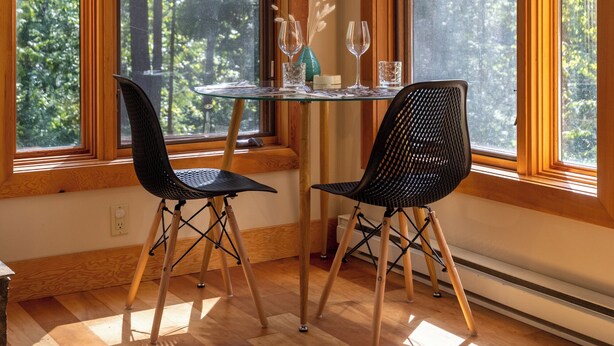 Une pièce dotée de grandes fenêtres, une table à manger avec des chaises, et une petite table sur laquelle se trouvent un vase et des verres.