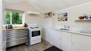 A compact kitchen with white cabinets, a stove, a sink, and a window with a view of greenery. - Okiwi Bay Cosy Cottage (Ōkiwi Bay)