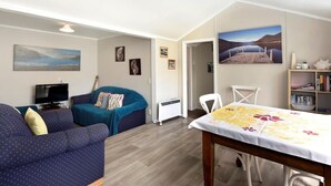 A cozy living room with a sofa, a TV, and a dining table with a tablecloth. - Okiwi Bay Cosy Cottage (Ōkiwi Bay)