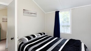 A bedroom with a bed, a window with curtains, and a wall-mounted air conditioning unit. - Okiwi Bay Cosy Cottage (Ōkiwi Bay)