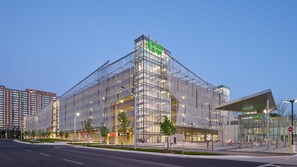A modern building with a glass facade, a green logo at the top, and a covered entrance area.