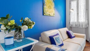 Habitación con pared azul, sofá blanco con cojines azules y morados, una mesa con un jarrón de flores y un cuadro.