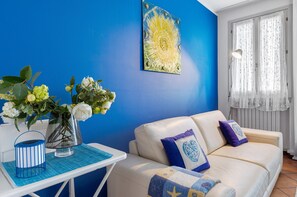 A room with a blue wall, a white sofa with blue and purple cushions, a table with a vase of flowers, and a painting.
