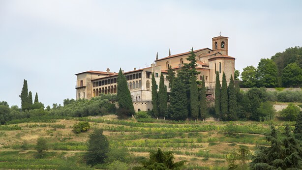 Un edificio grande con varios arcos y una torre del campanario, rodeado de árboles y colinas.
