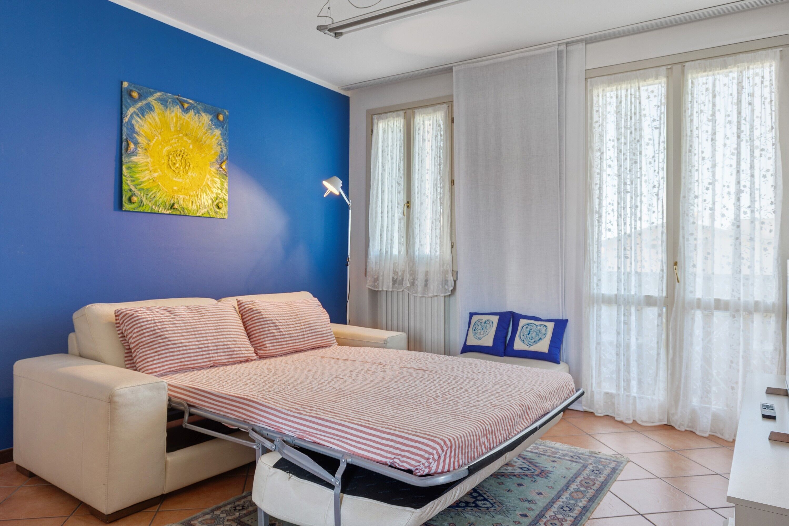 A bedroom with a bed, a sofa, a window with curtains, and a wall painting.