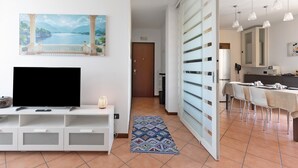 Una sala de estar moderna con un televisor de pantalla plana, un cuadro de una escena costera, un comedor con mesa y sillas, y una cocina con armarios y un refrigerador.