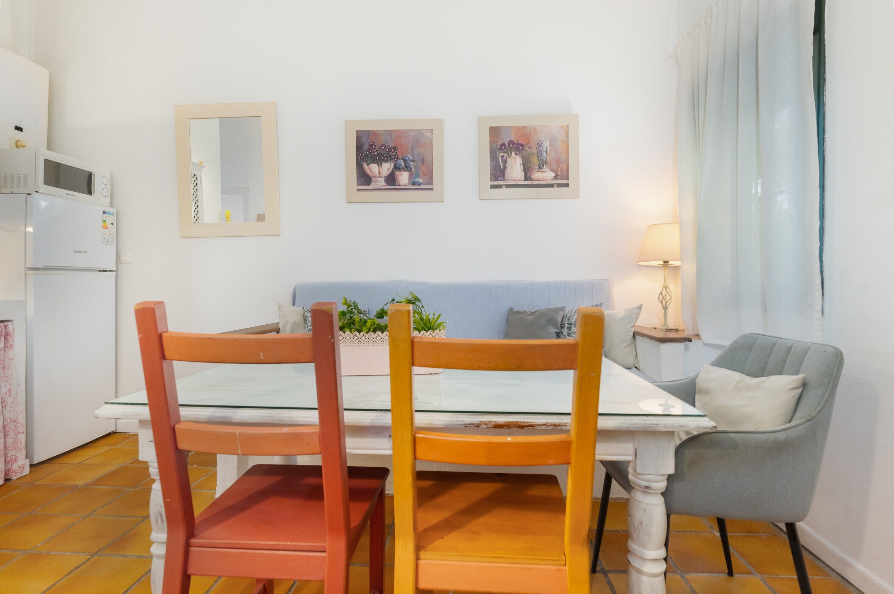 Un comedor con una mesa blanca, dos sillas de madera, un sillón gris, una mesita auxiliar con una lámpara y dos cuadros enmarcados en la pared.