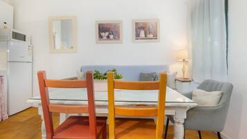 Un comedor con una mesa blanca, dos sillas de madera, un sillón gris, una mesita auxiliar con una lámpara y dos cuadros enmarcados en la pared.