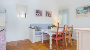 A small, clean room with a white table, two chairs, a sofa, and a mirror. There are two framed pictures on the wall and a window with curtains.