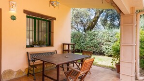 A patio with a wooden table and chairs, a window with green shutters, and a view of a garden.
