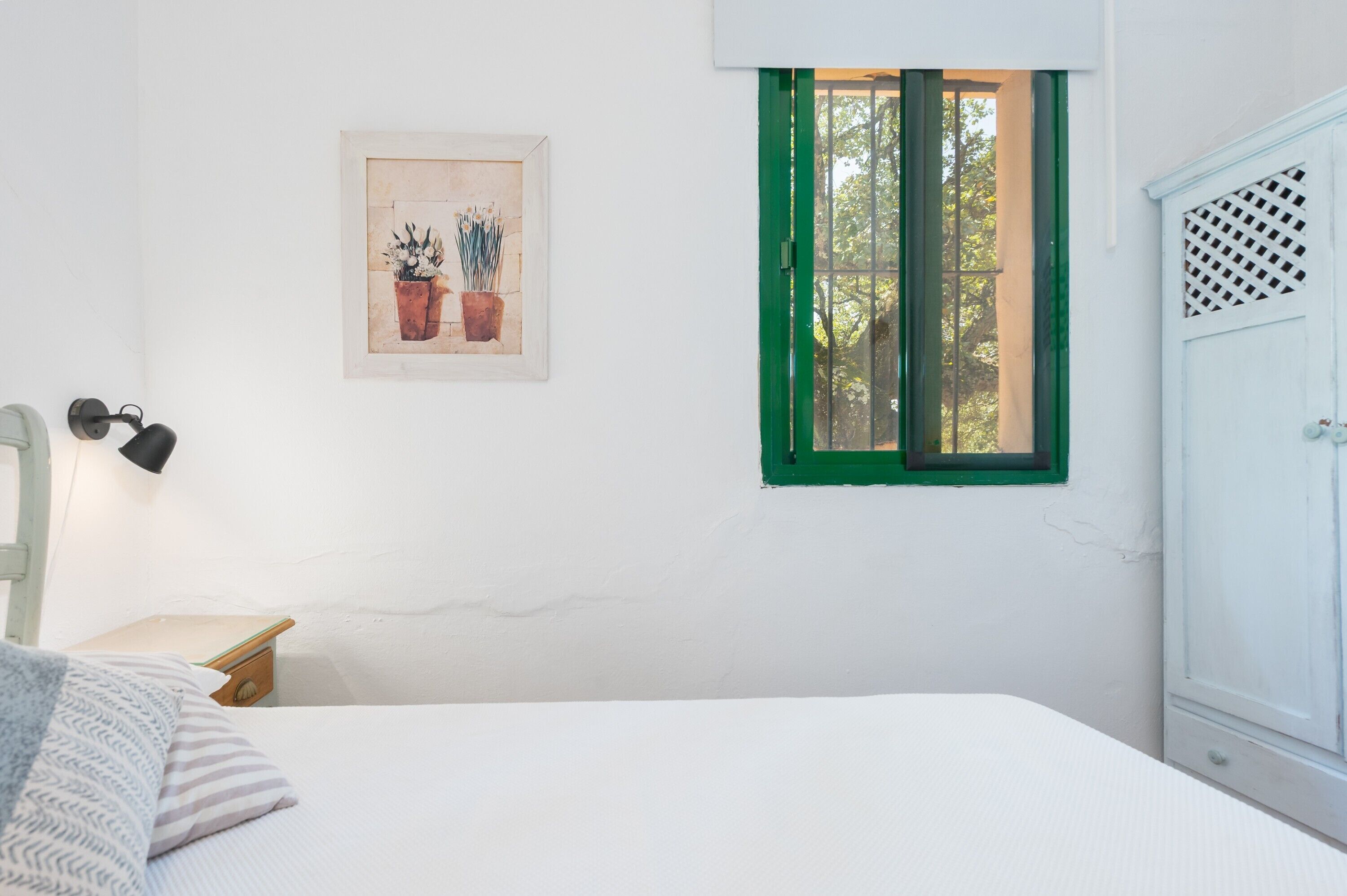 Une chambre à coucher comprenant un lit, une table de chevet, une lampe, une fenêtre avec des volets verts, un tableau accroché au mur et une armoire.