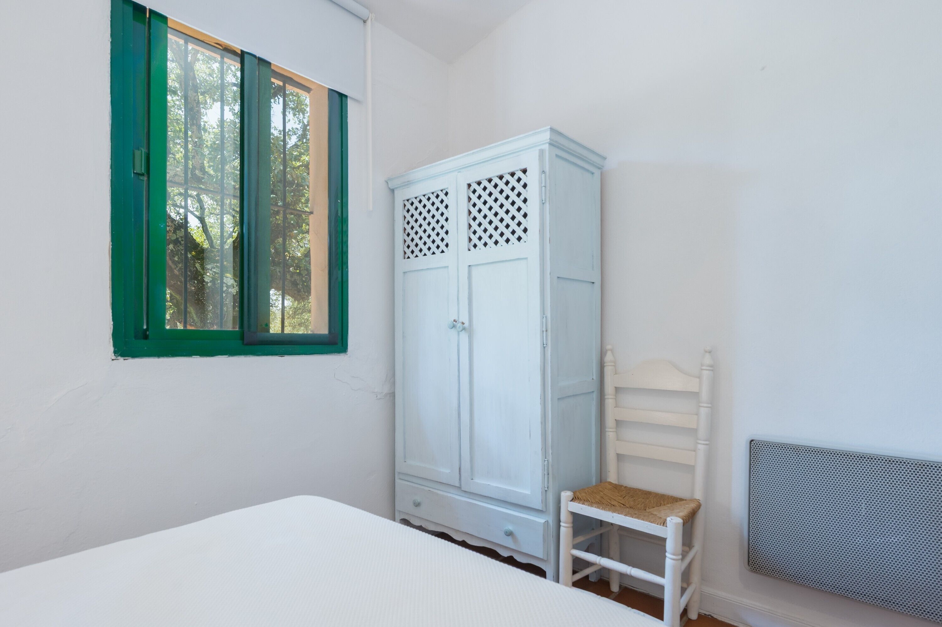 Une pièce avec un lit, une armoire blanche, une fenêtre avec des volets verts et un radiateur.