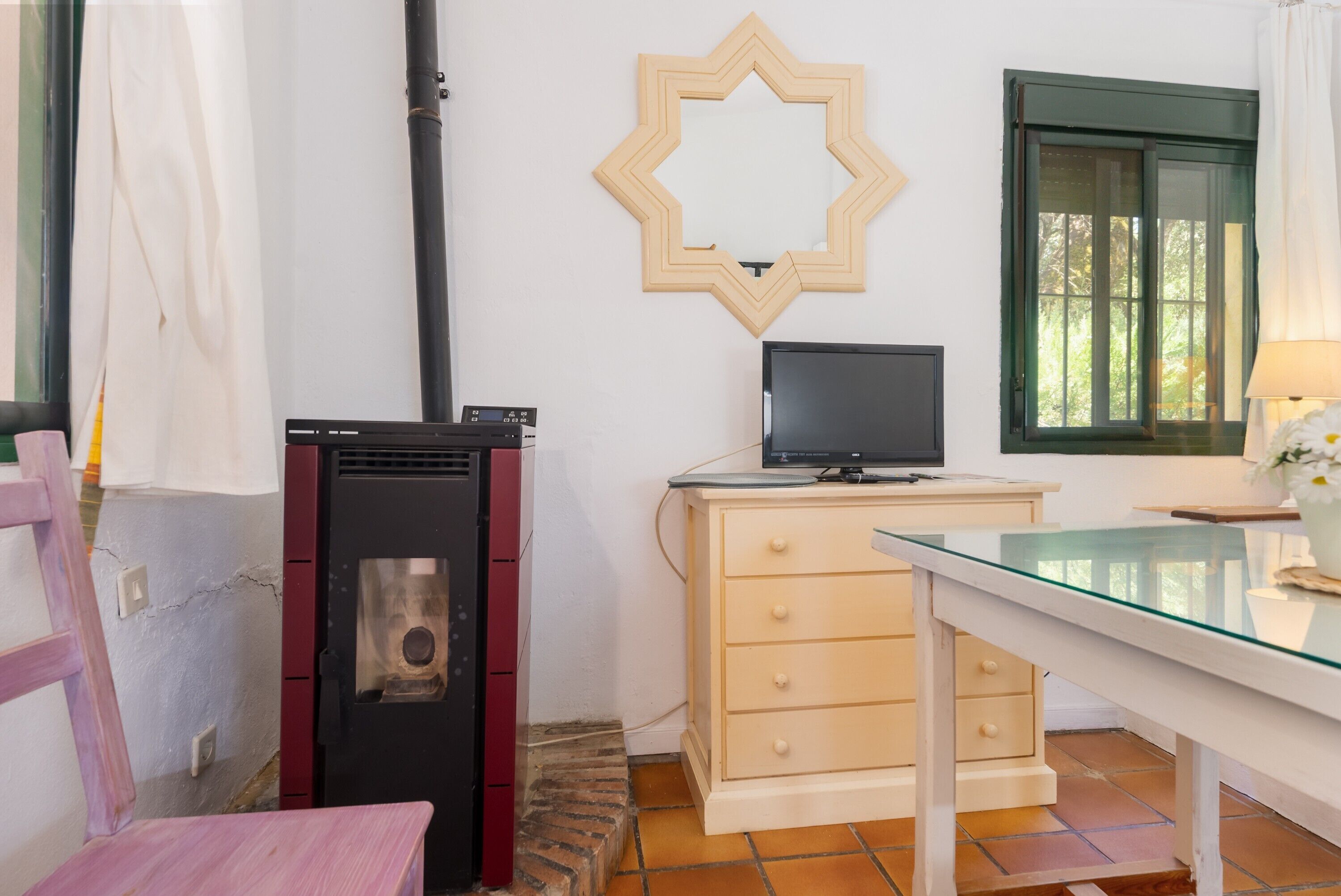 A room with a wood-burning stove, a TV on a dresser, a glass table, and a window with a view of trees.