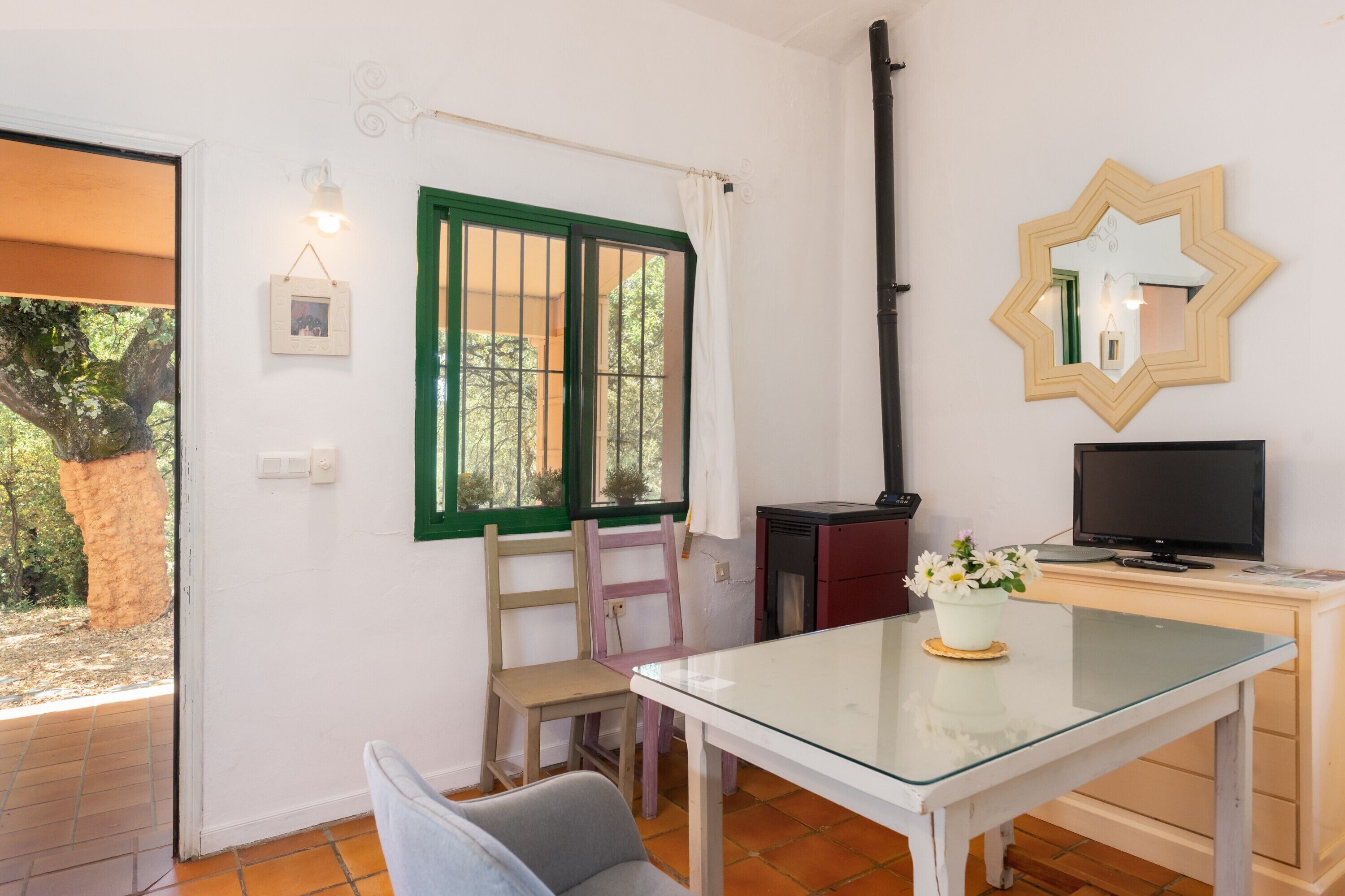 Una habitación con una mesa de vidrio, televisor, silla, escritorio, espejo y ventana con contraventanas verdes.