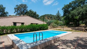 A swimming pool with two ladders, surrounded by a paved area with red tiles, and a building with a sloped roof in the background. - Rural House El Chopo with Mountain Views, Terrace, Shared Pool, and Parking (Alajar)