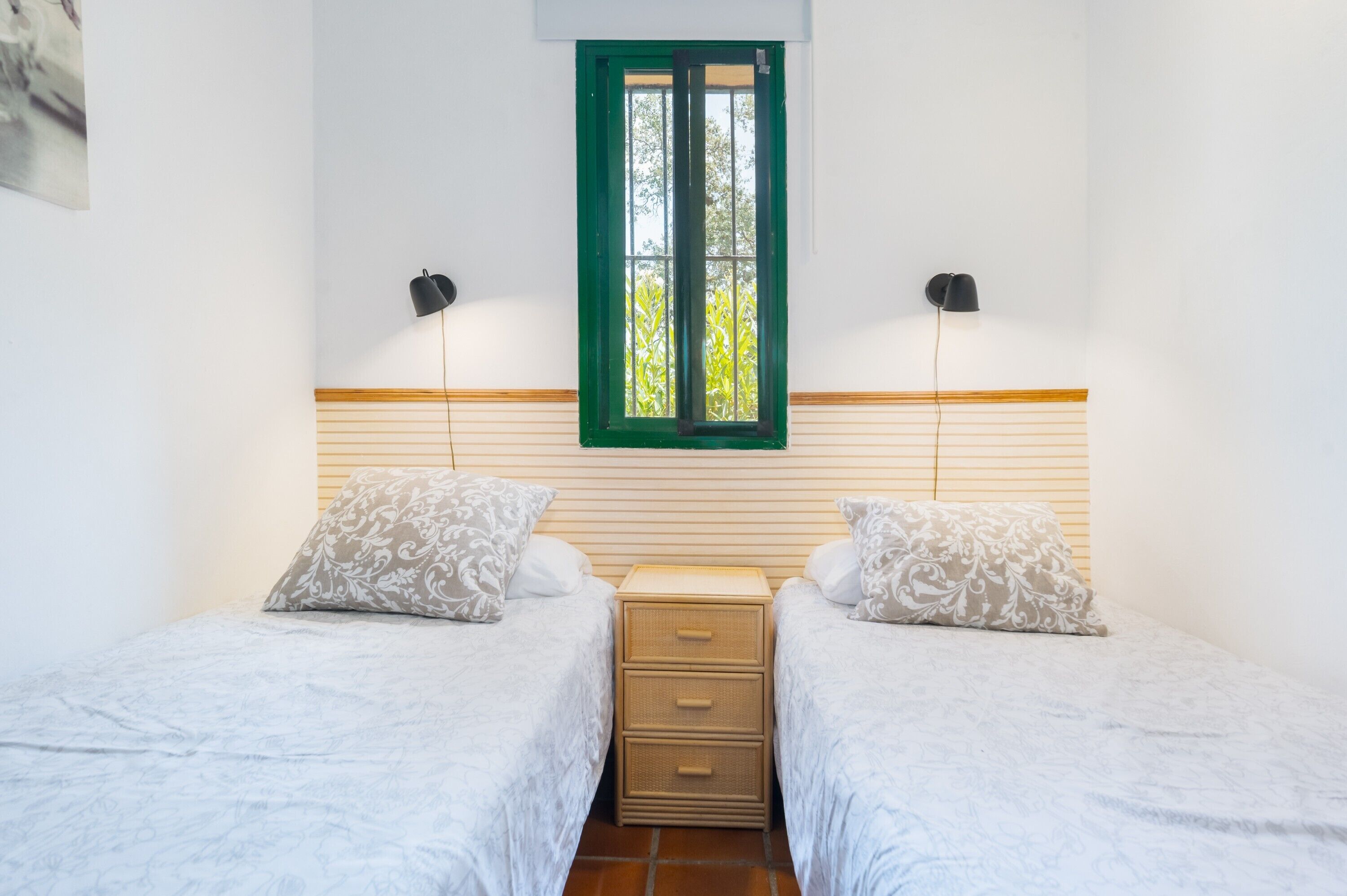 A bedroom with two single beds, a wooden nightstand, a green-framed window, and two wall-mounted lamps.