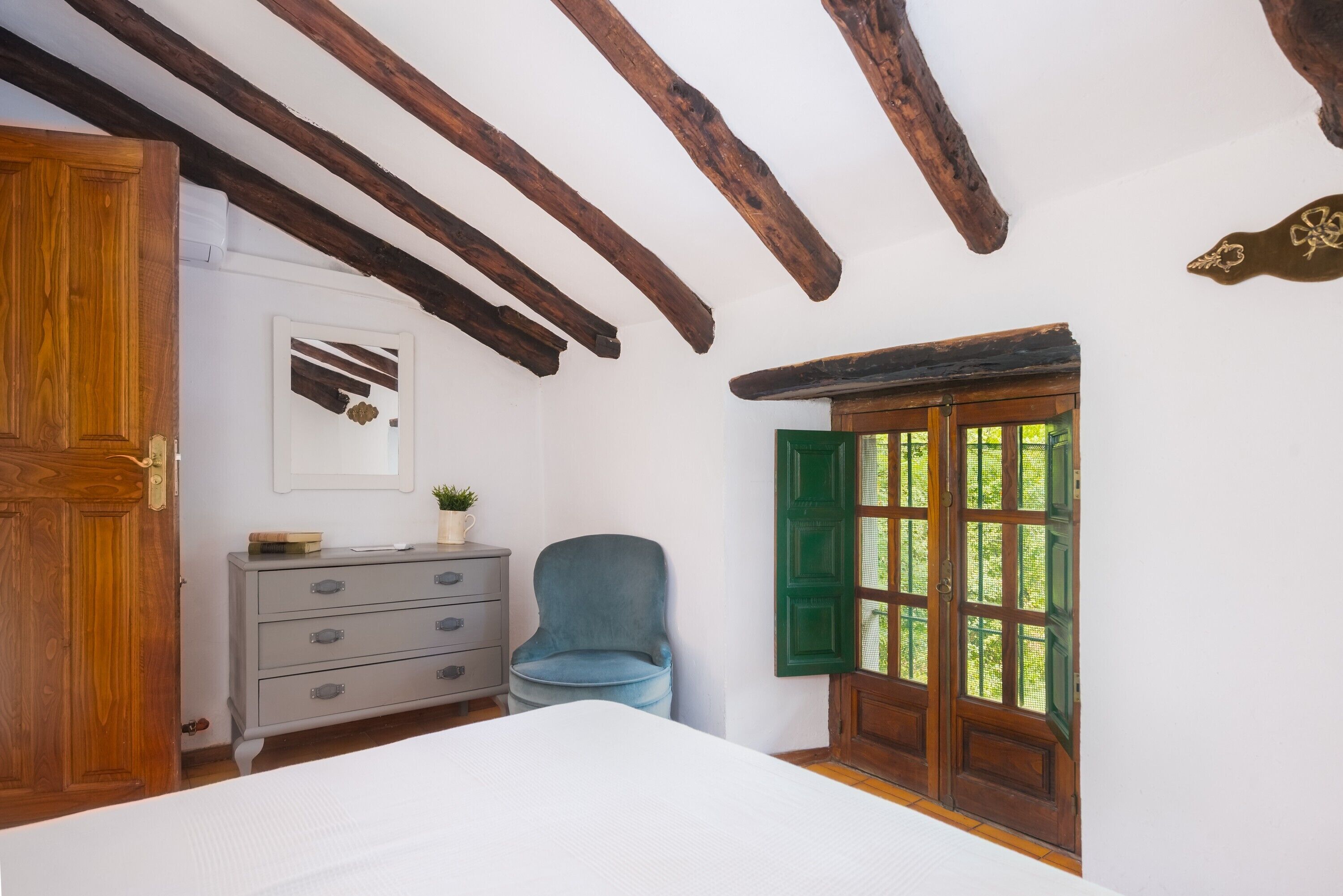 A bedroom with a wooden ceiling, a bed, a dresser, a chair, and a window with green shutters.
