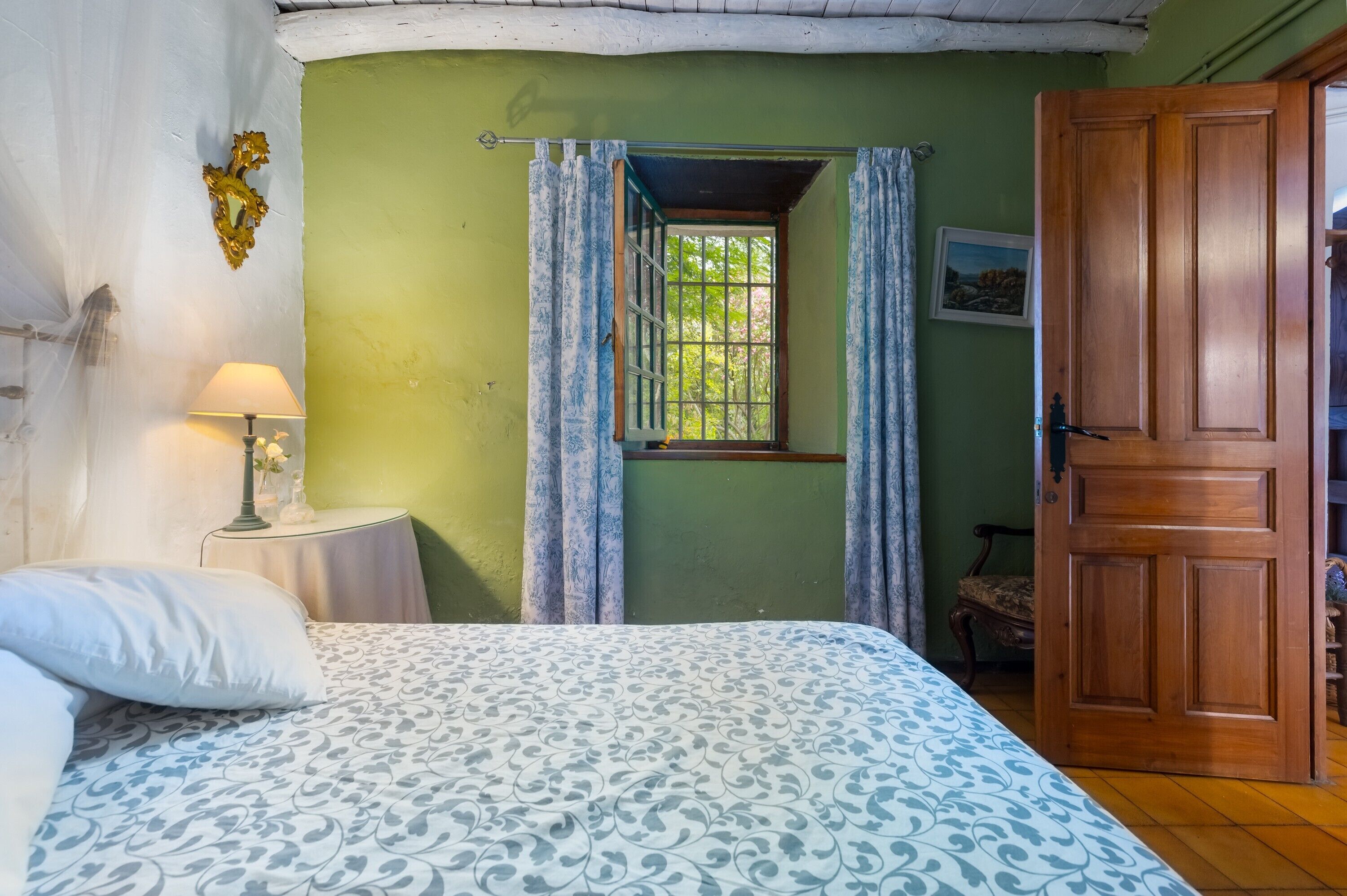 Een slaapkamer met een bed, een nachtkastje met een lamp, een raam met gordijnen, en een houten deur.