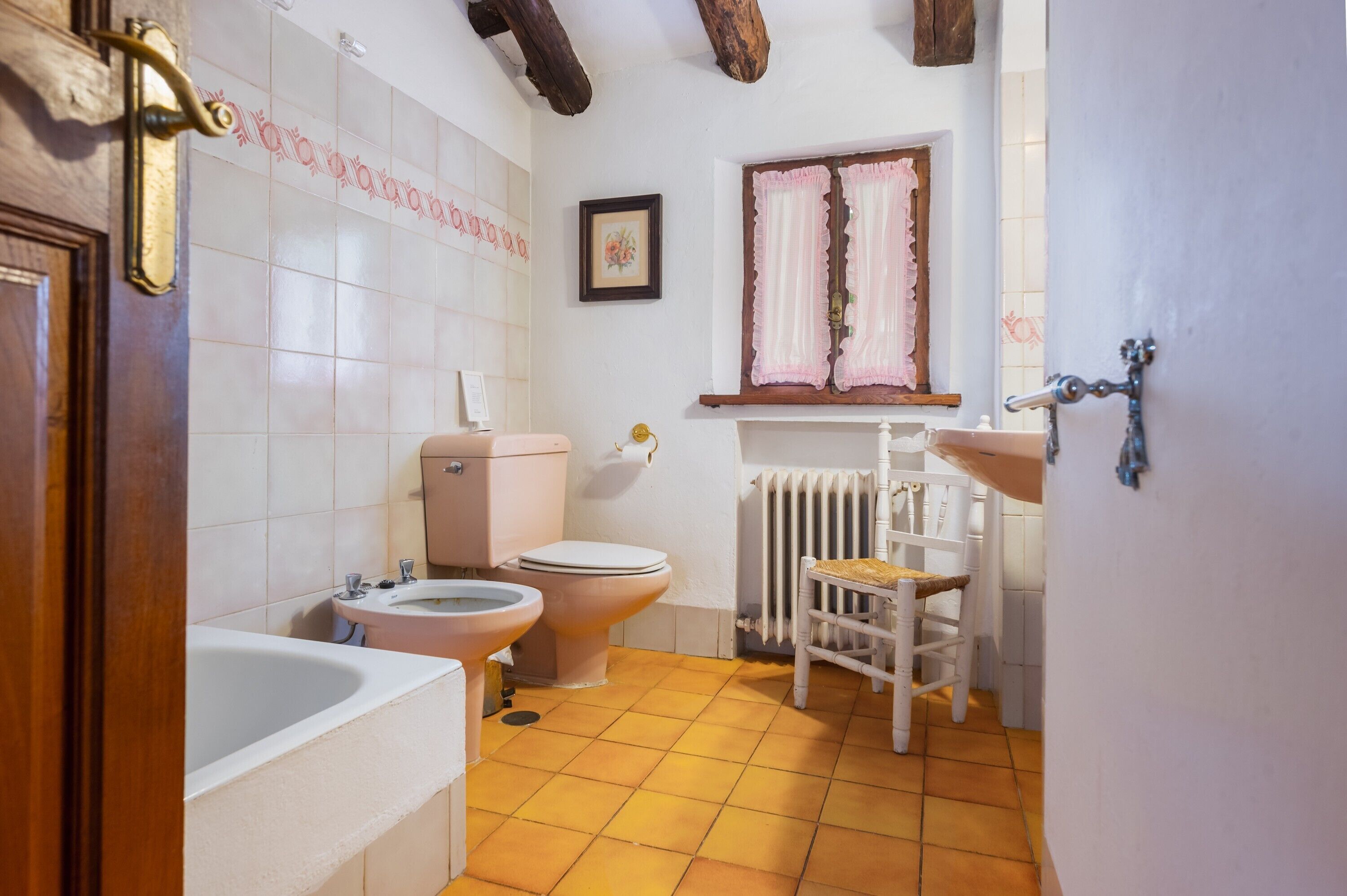 A bathroom with two toilets, a sink, a bathtub, a radiator, and a wooden chair.