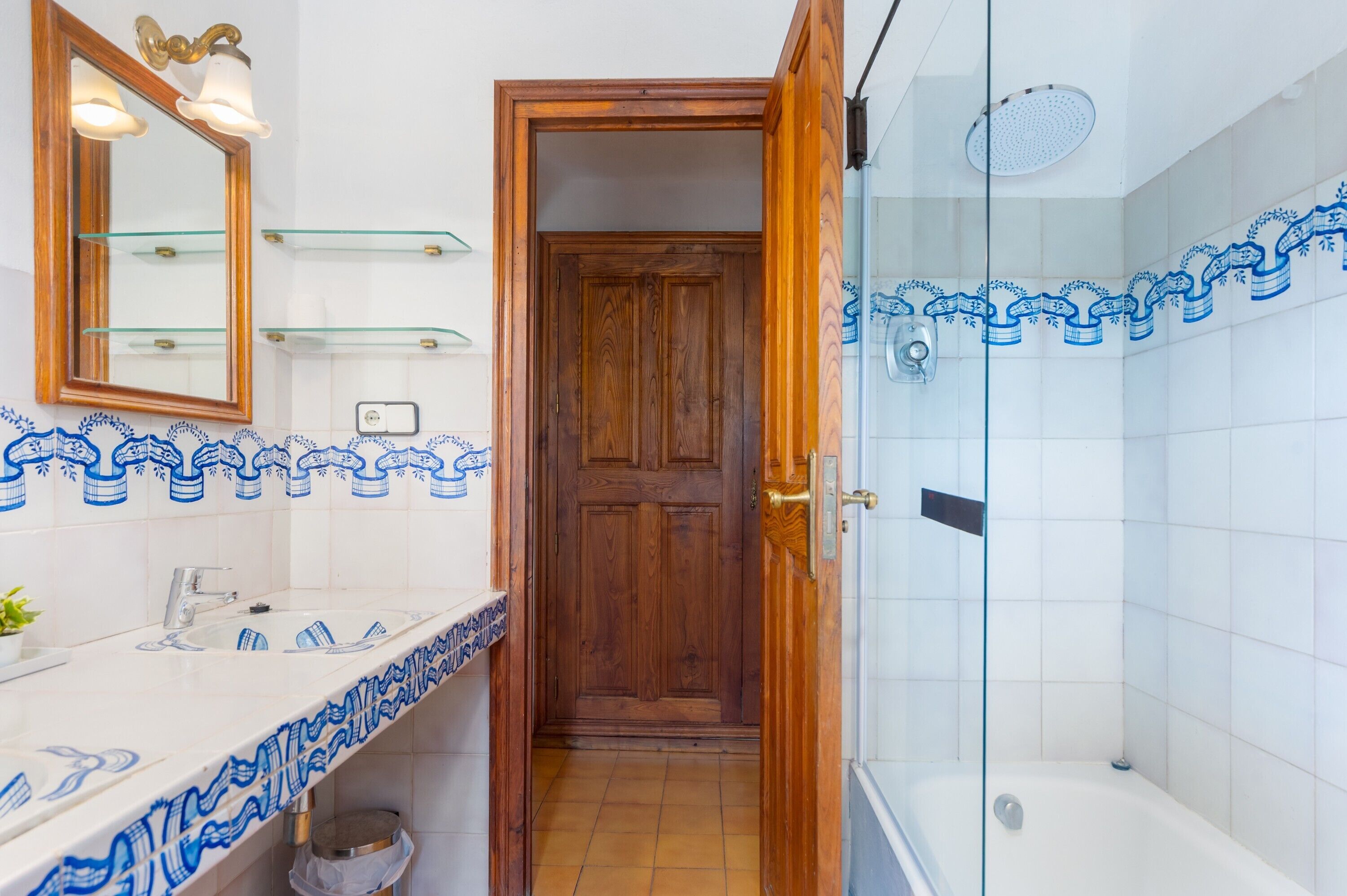 A bathroom with a wooden door, a glass shower enclosure, and a white sink with blue patterned tiles.