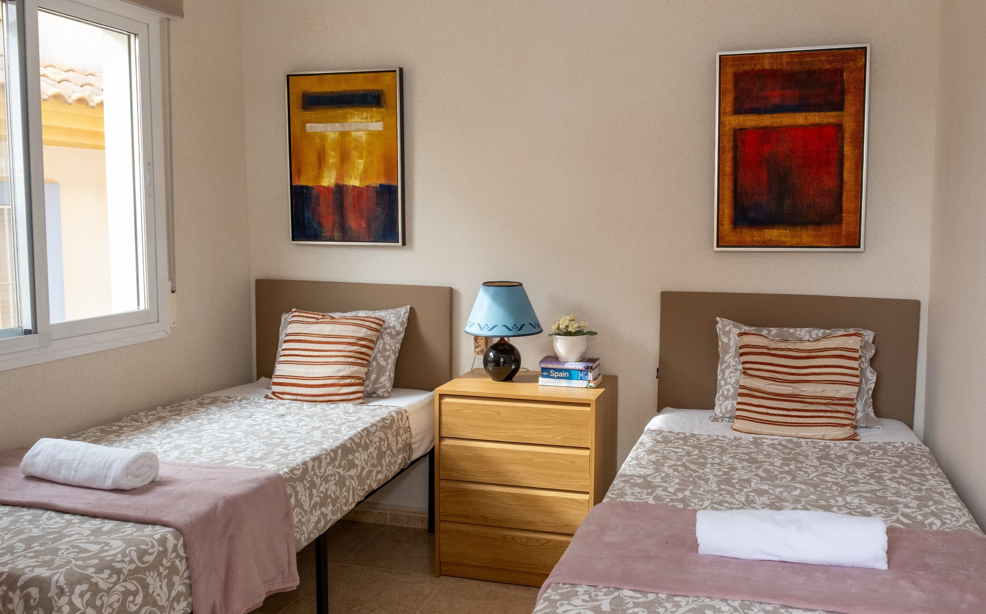 A hotel room with two single beds, a nightstand, a lamp, a potted plant, and two abstract paintings on the wall.
