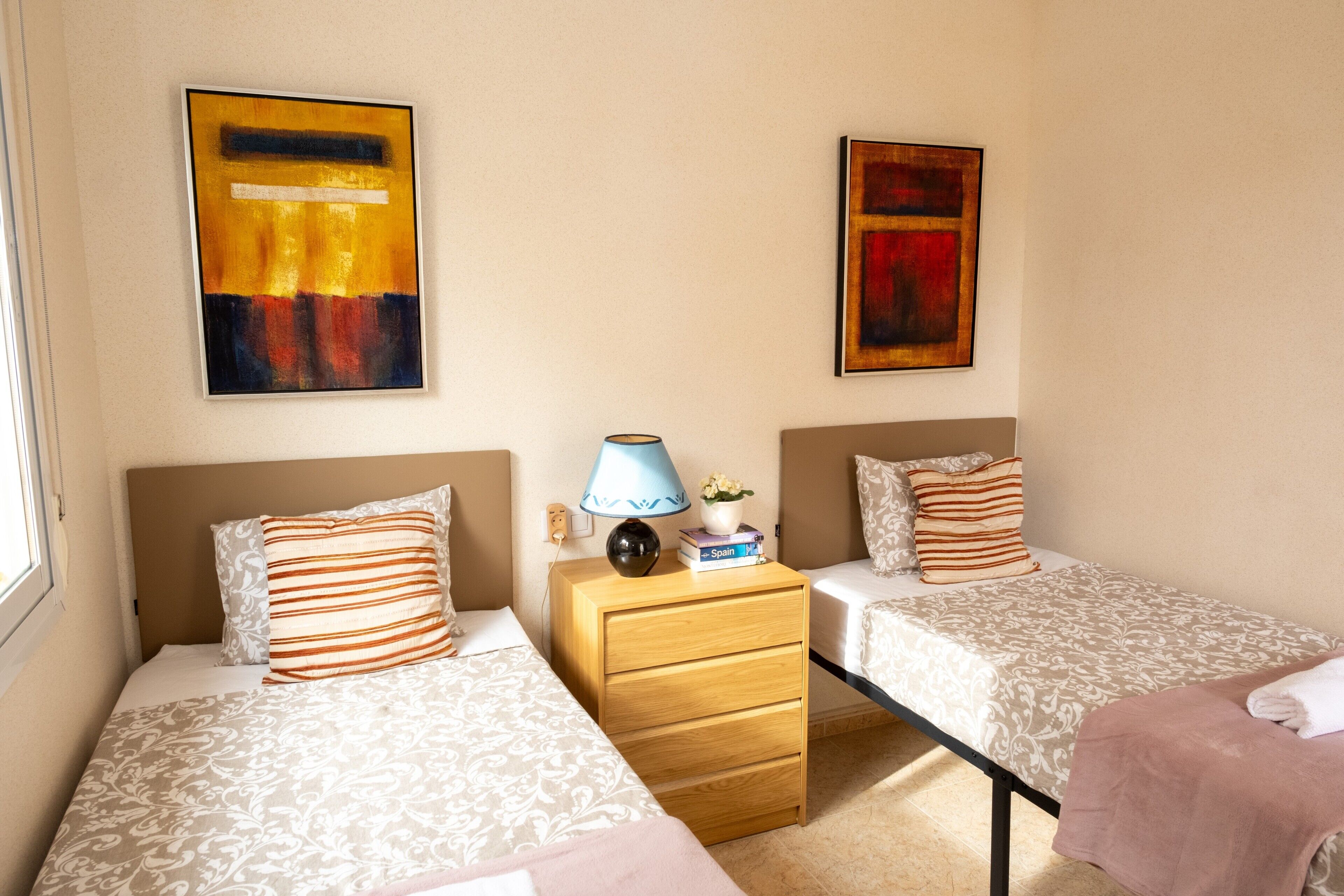 A hotel room with two beds, a nightstand, a lamp, and two paintings on the wall.