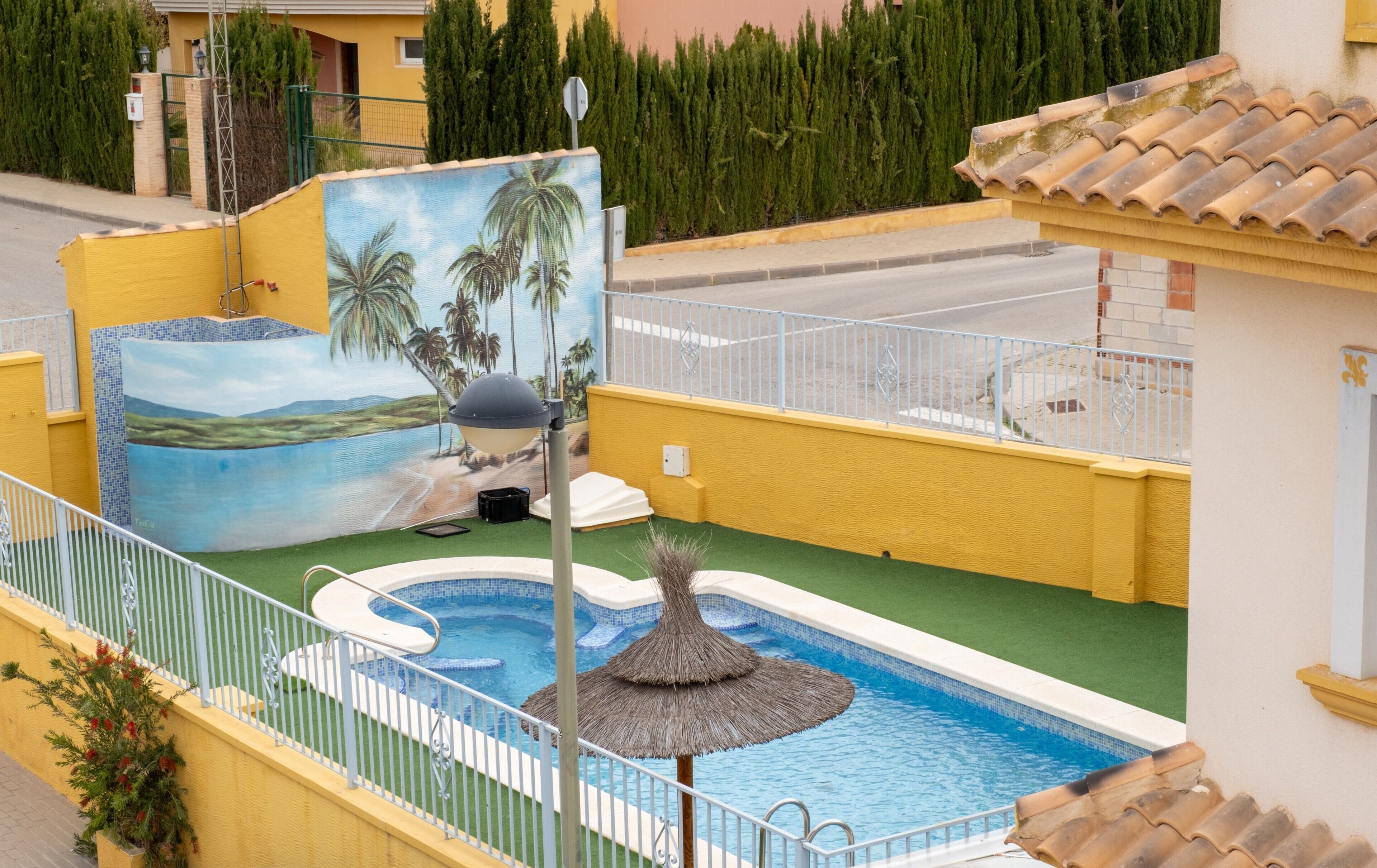 A pool with a tropical mural, a thatched umbrella, and a metal fence.