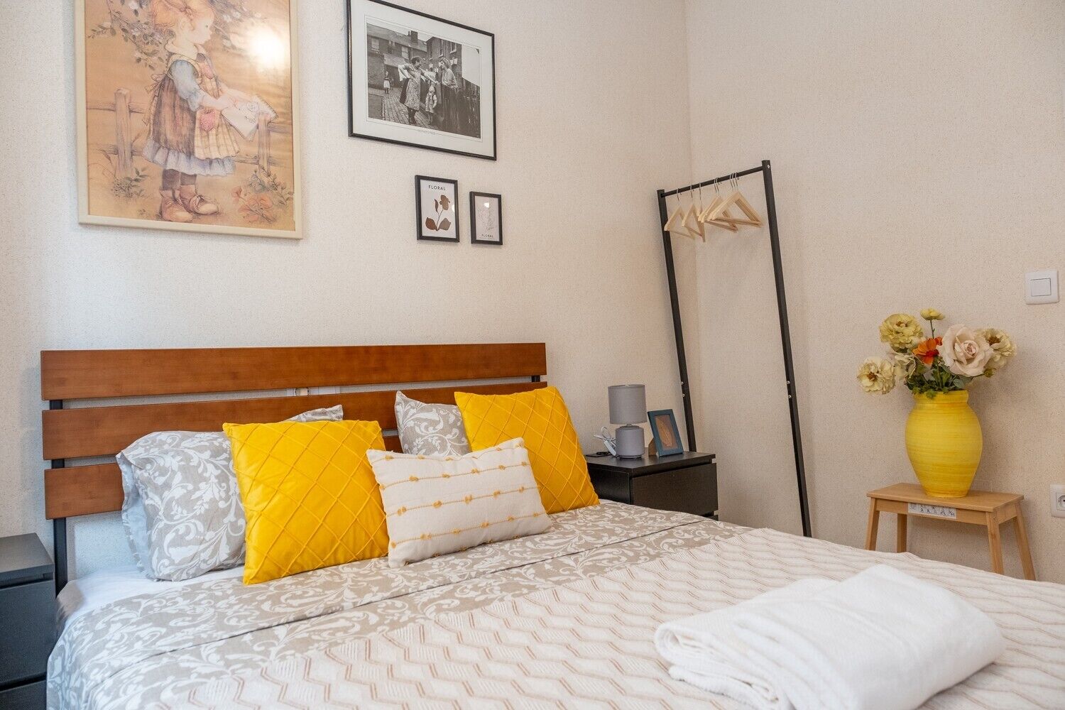 Una cama bien hecha con cabecera de madera, una mesita de noche con lámpara, un espejo y un florero con flores sobre una pequeña mesa.
