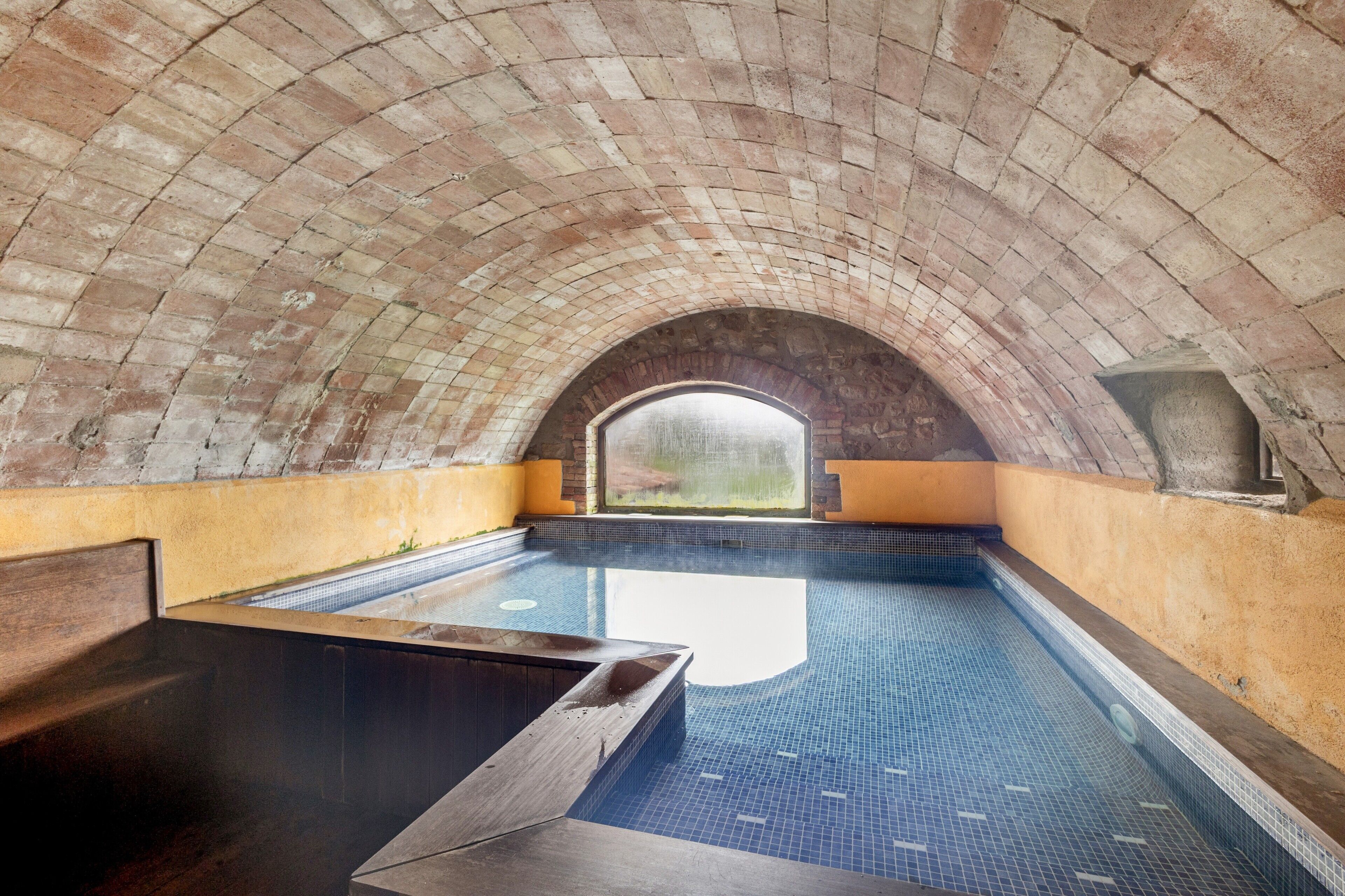 Una piscina interior con suelo de baldosas, un banco de madera y un techo abovedado de ladrillo.
