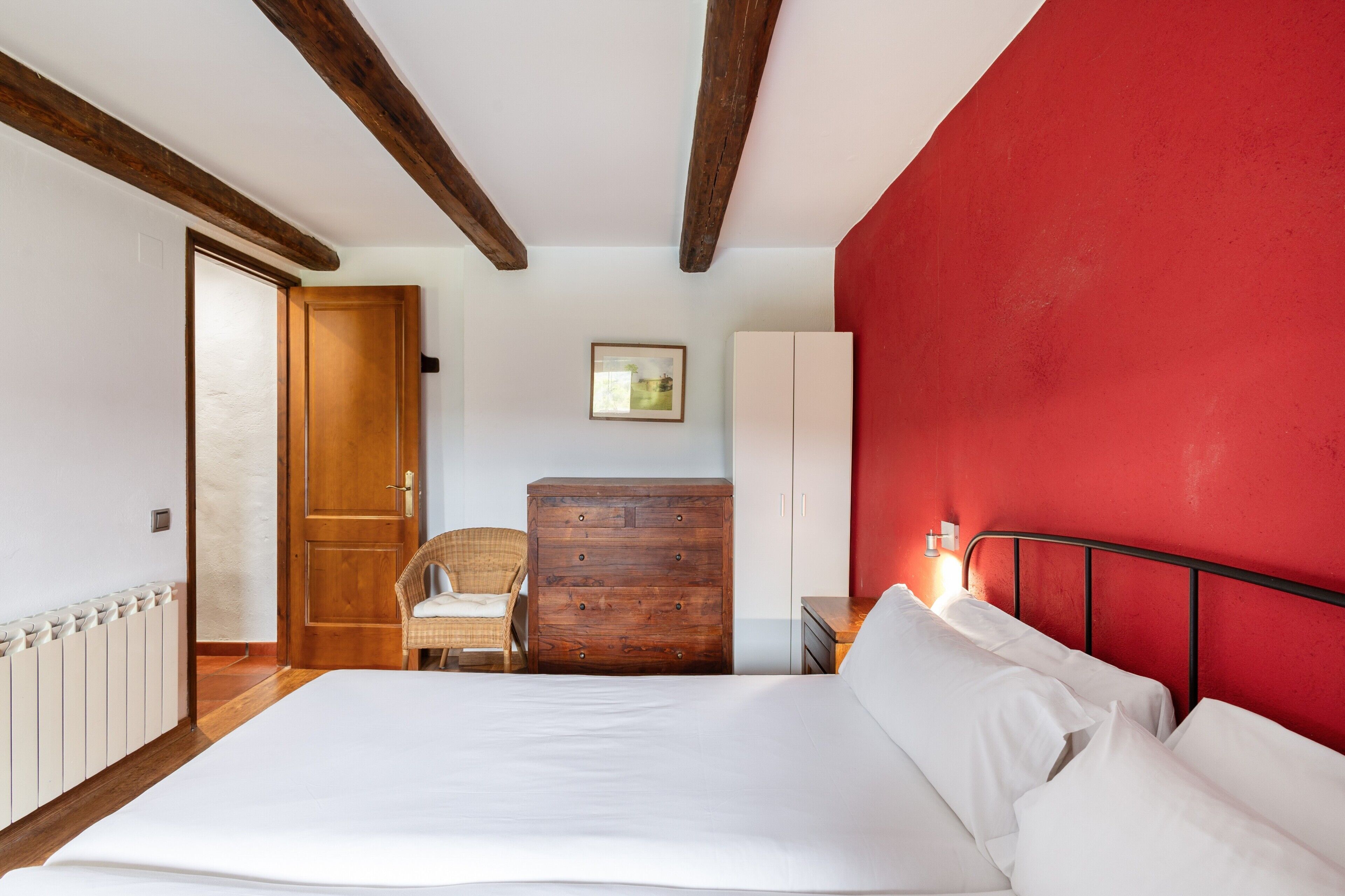 Ein Schlafzimmer mit Bett, Holzkommode, Stuhl und einer roten Akzentwand.