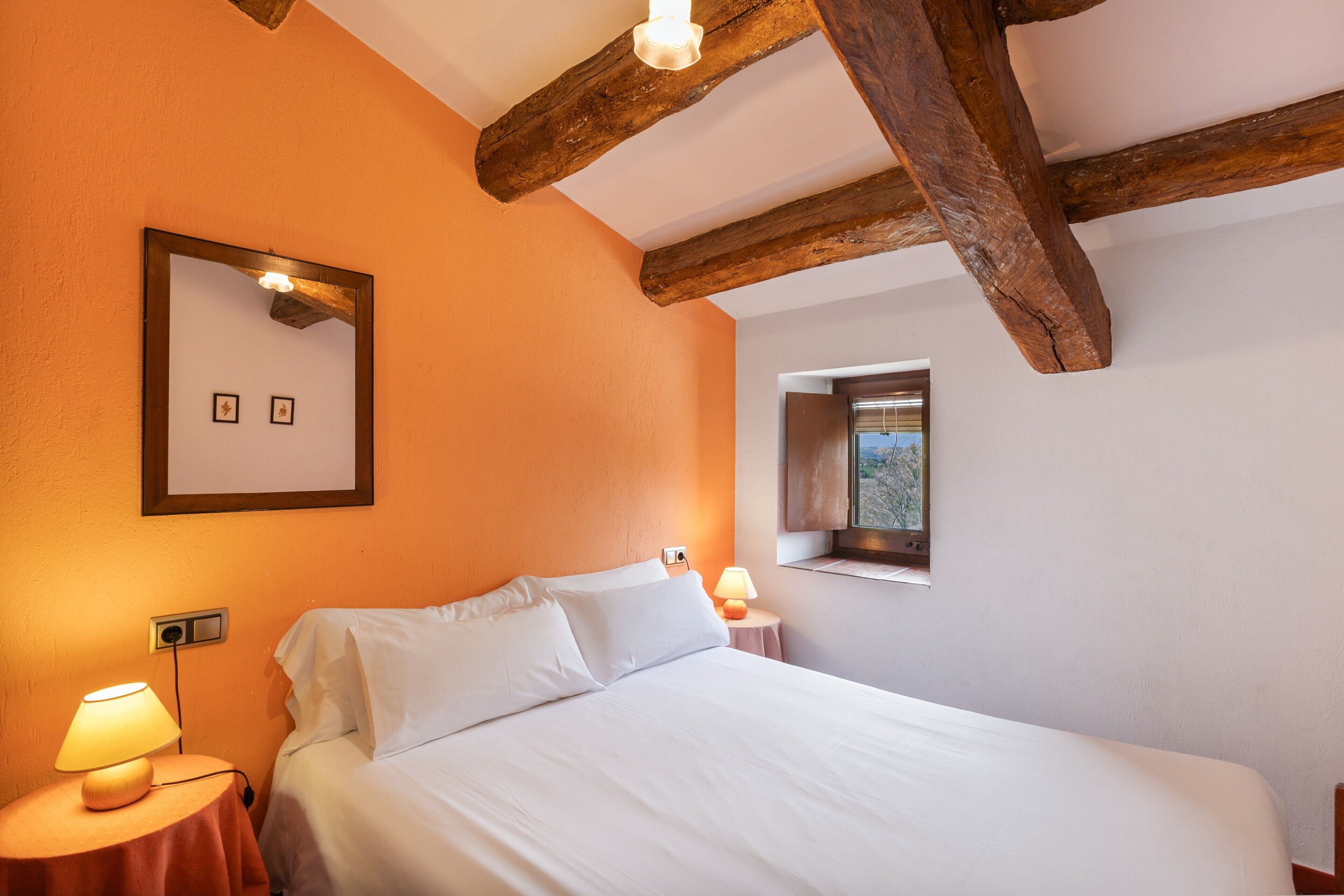 Un dormitorio con cama, lámparas de noche, ventana y vigas de madera a la vista.