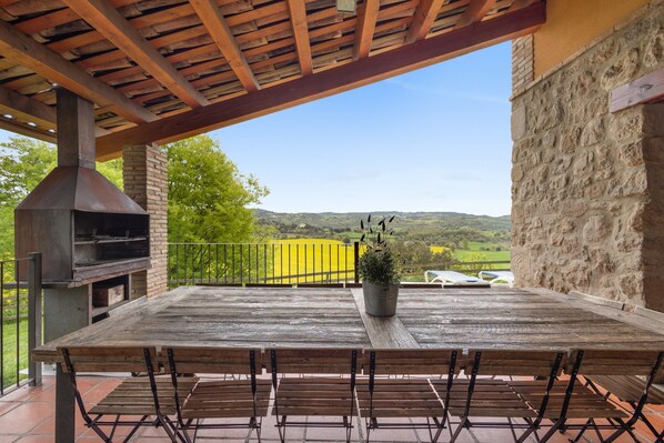Un patio exterior con mesa y sillas de madera, una chimenea de ladrillo y vistas a colinas verdes.