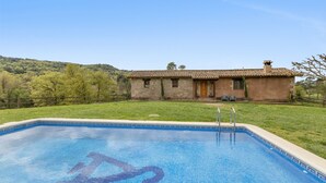 Una piscina con borde de azulejos, un edificio de piedra con techo de tejas y una zona verde con árboles al fondo.