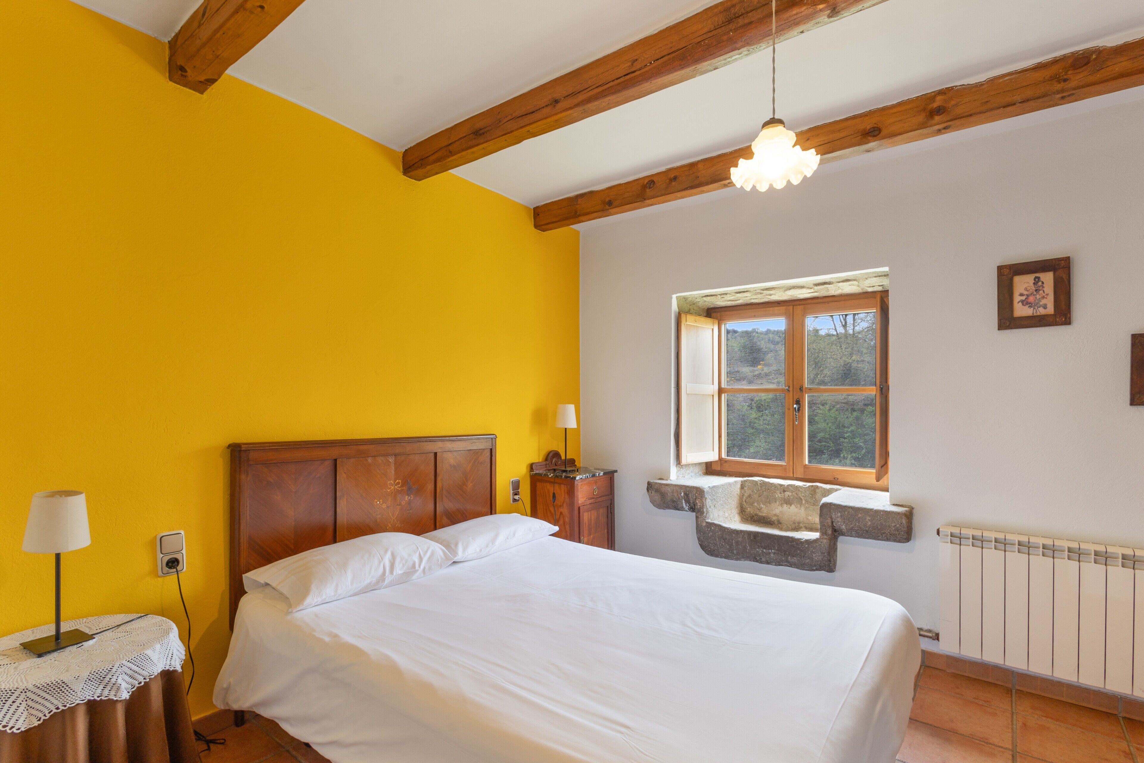 Une chambre avec un plafond en bois, un lit recouvert de draps blancs, une table de chevet avec une lampe, une fenêtre dotée d’un rebord en pierre et un radiateur.