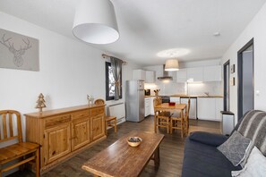 Una cocina moderna con armarios blancos, un comedor con mesa y sillas de madera, un sofá con una manta estampada y un banco de madera con un cuenco.