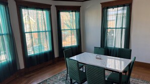 Un comedor con una mesa blanca y sillas verdes, ventanas grandes con cortinas y una alfombra estampada sobre el piso de madera.