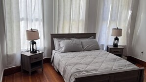 Un dormitorio con suelo de madera, una cama con colcha gris, dos mesitas de noche con lámparas y cortinas de encaje con lazos.