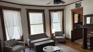 Una sala de estar con un sofá gris, dos sillones, una mesa de centro y un ventilador de techo. Hay ventanas amplias con cortinas traslúcidas, una zona de cocina con armarios de madera y una frase motivadora en la pared.