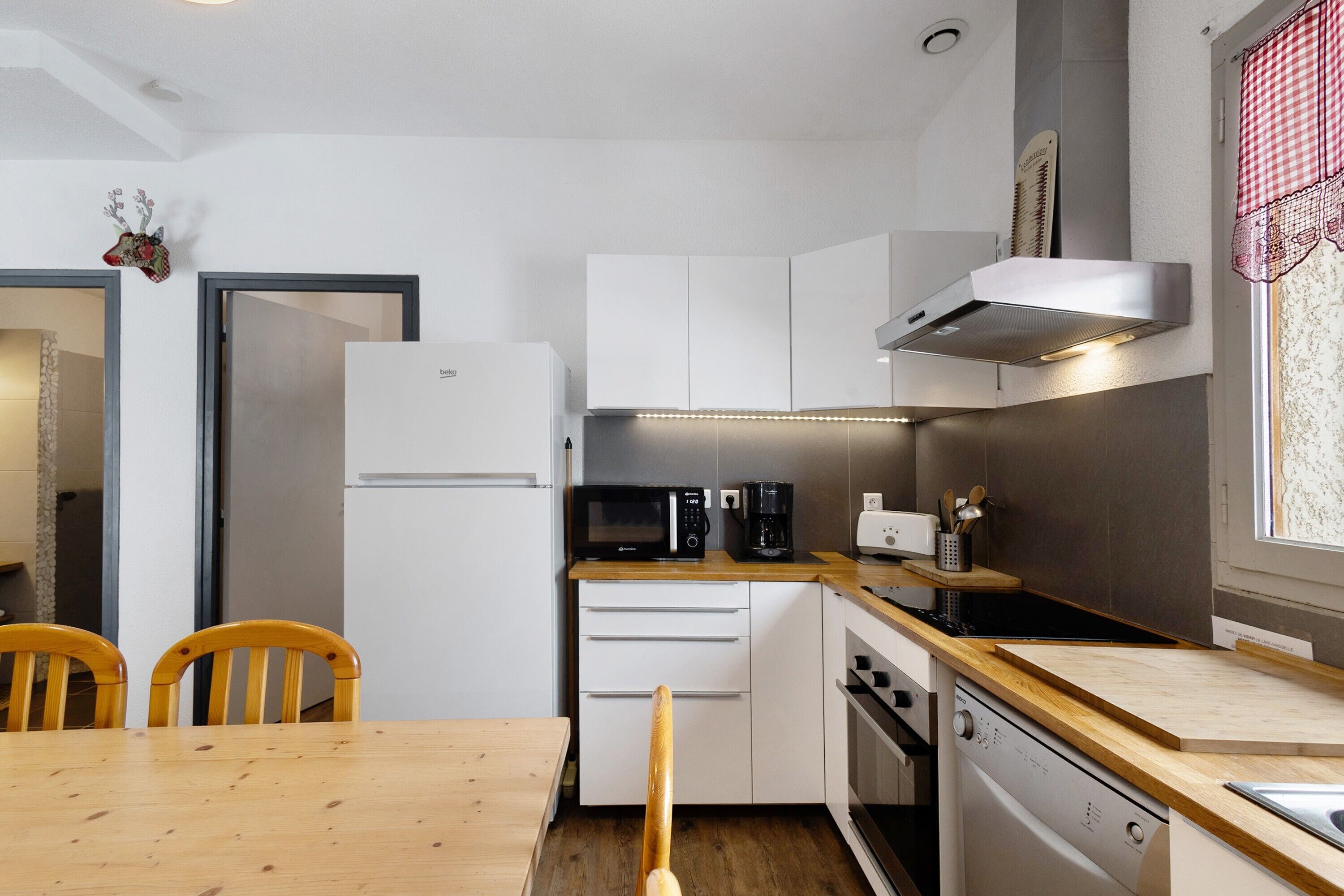 Una cucina compatta con mobili bianchi, un forno a microonde, una macchina per caffè e una lavastoviglie. C'è un tavolo da pranzo in legno con due sedie.