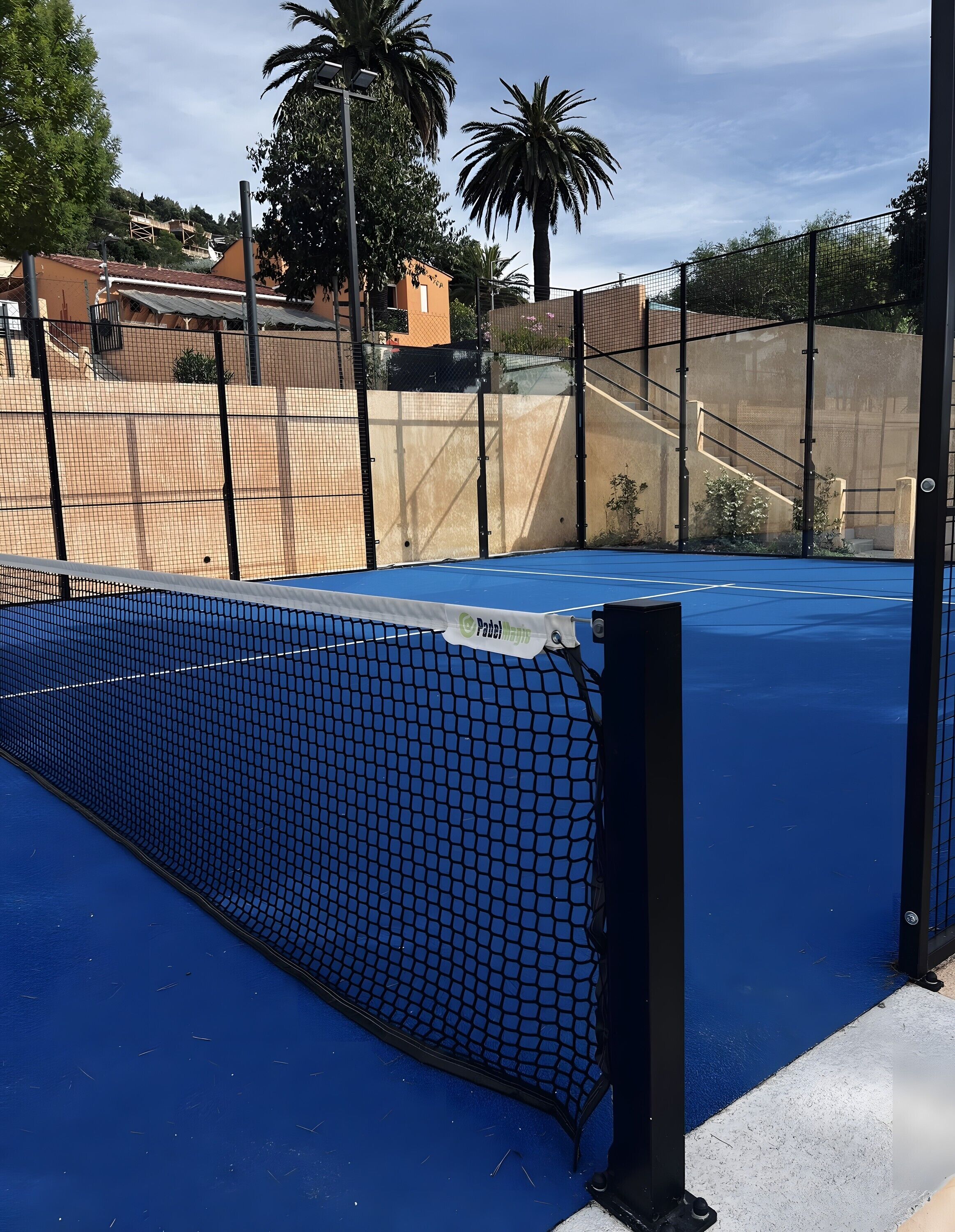 Una cancha de tenis con red, rodeada por una valla y palmeras.