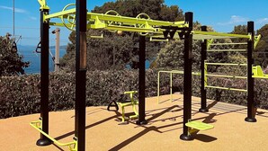 Een buitenoefenruimte met diverse fitnessapparaten, waaronder een optrekstang, een bank en een beenspersapparaat.