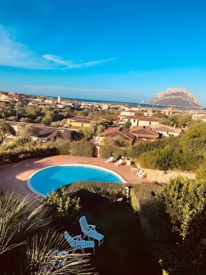 Una piscina con sillones de descanso, montañas al fondo y un pueblo costero.