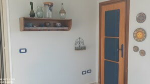 Quarto com porta de madeira, prateleira com objetos decorativos, interruptores de luz fixados na parede e pratos decorativos.